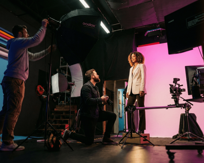A photographer kneels in front of a model in a studio, while another person adjusts a large softbox light.
