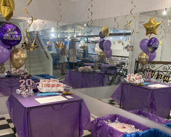 A decorated party space with two tables covered in purple tablecloths, surrounded by balloons and stars, with one table displaying gifts and the other containing ice and beverages.