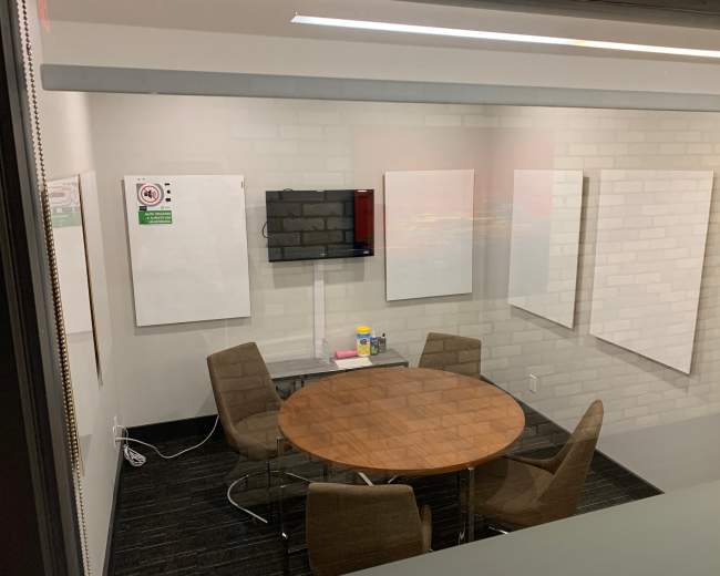 The image shows a conference room with a round wooden table surrounded by four chairs, a television mounted on the wall, and several blank whiteboards.