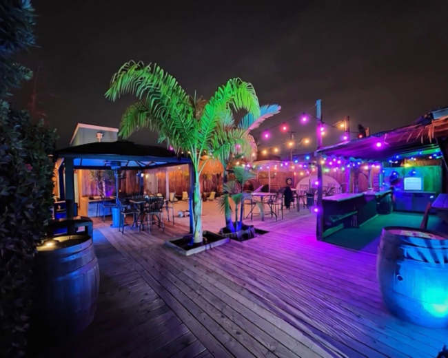 The image shows an outdoor patio area illuminated by colorful string lights, featuring a palm tree, seating arrangements, and wooden decking.