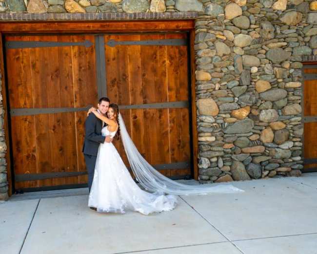 A couple embraces in formal attire in front of a stone wall with large wooden doors.