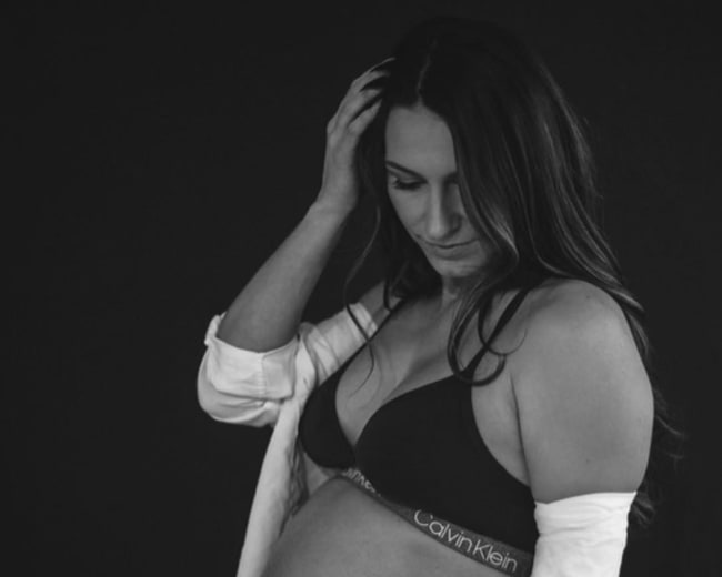 A woman in a black bra and white shirt gently holds her pregnant belly while looking down.