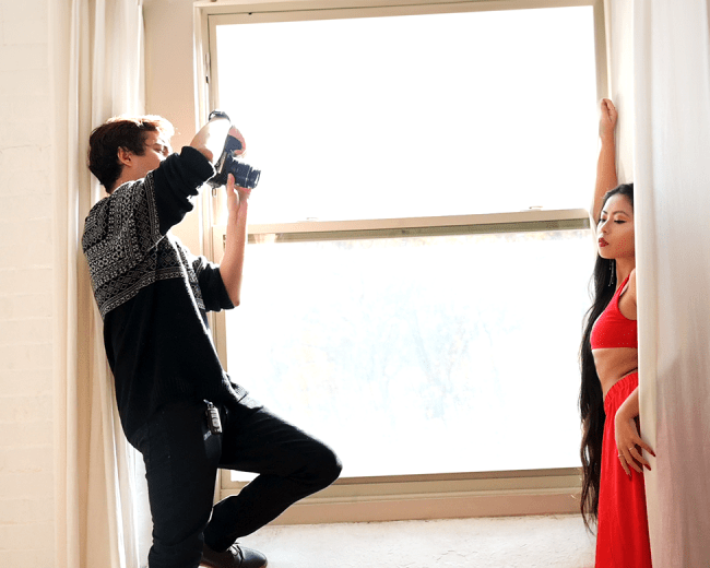 A photographer captures an artistic image of a model posing by a large window draped with sheer curtains.