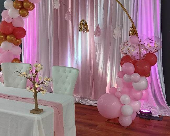 A decorated event area features a white table with a floral centerpiece and an arch adorned with pink, gold, and white balloons, set against a backdrop of sheer pink drapery.