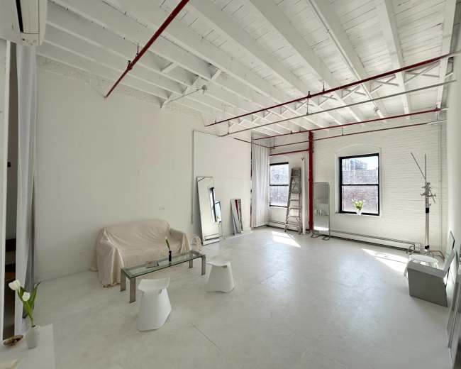 The image shows a spacious, empty studio with white walls and a high ceiling, featuring a sofa covered with a sheet, a glass coffee table, and minimal furnishings.