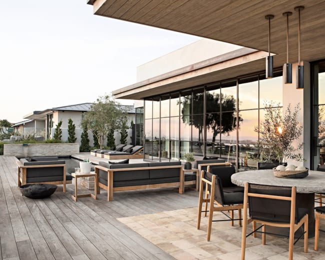A modern outdoor patio features wooden decking, a circular stone dining table surrounded by chairs, and large glass windows reflecting the landscape.