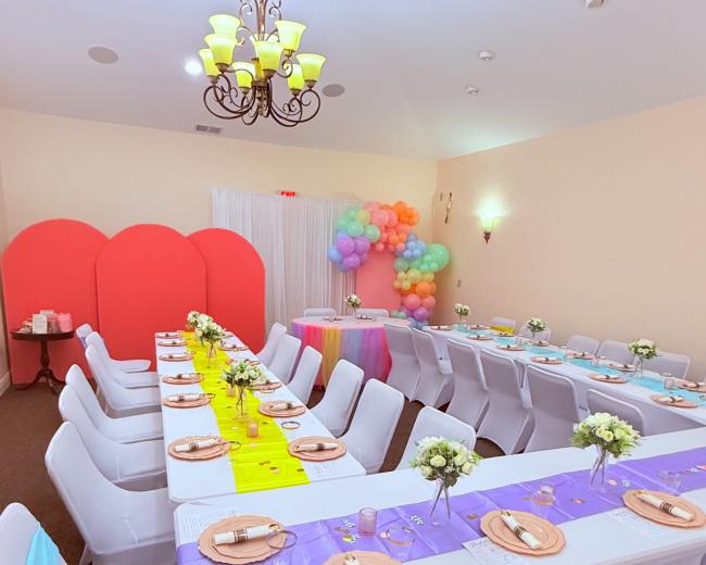 A decorated event space features long tables set with colorful tablecloths and centerpieces, surrounded by white chairs adorned with ribbons.