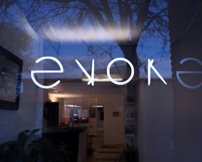The image shows the word "evoke" displayed in a mirrored format on a glass window, reflecting trees and a building interior.