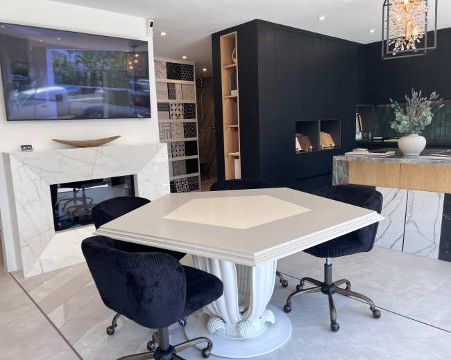 The image shows a modern interior space featuring a square table surrounded by black upholstered chairs, a television mounted on the wall, and a fireplace set into a marble wall.