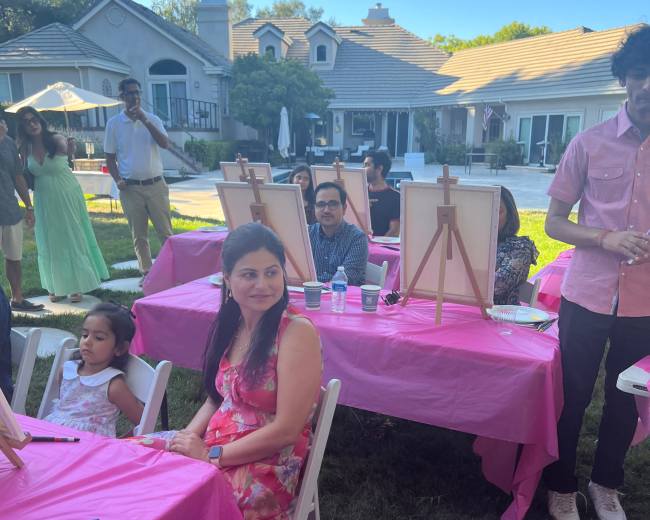 A group of people gathers around pink tables set up for a painting event in a backyard with a house in the background.