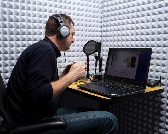 A person wearing headphones is sitting at a desk with a laptop and a microphone in a soundproof room.