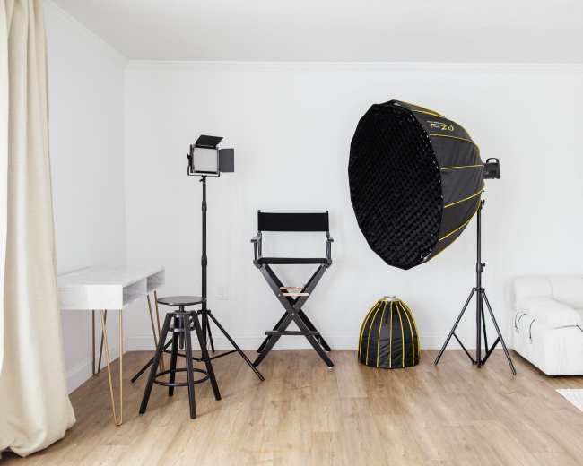 The scene shows a minimalist studio setting with a director's chair, lighting equipment, a large softbox, and a white sofa against a light-colored wall.