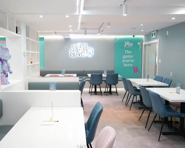 The image shows a brightly lit, modern café interior featuring tables and chairs arranged neatly, with vibrant decor and a neon sign on the wall.