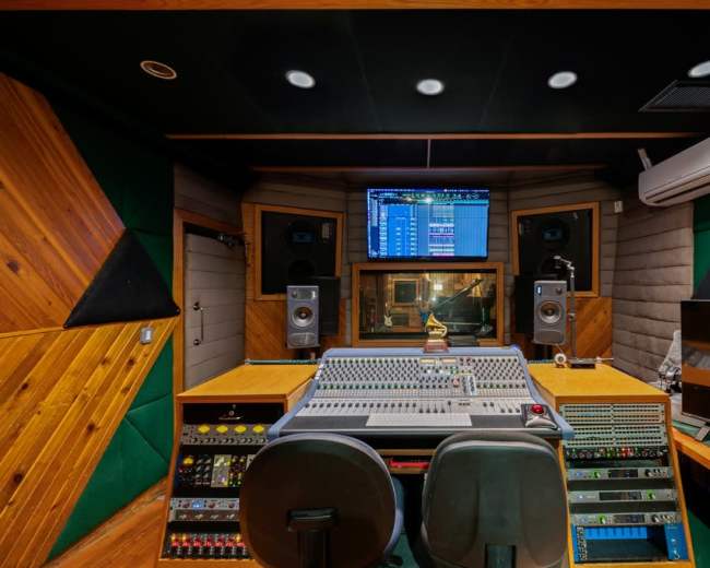 The image shows a sound studio with a large mixing console, speakers, and a monitor displaying audio tracks.