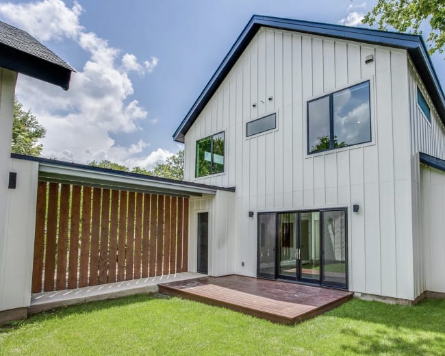 A modern two-story house features white vertical siding, large windows, and a wooden deck in the backyard.