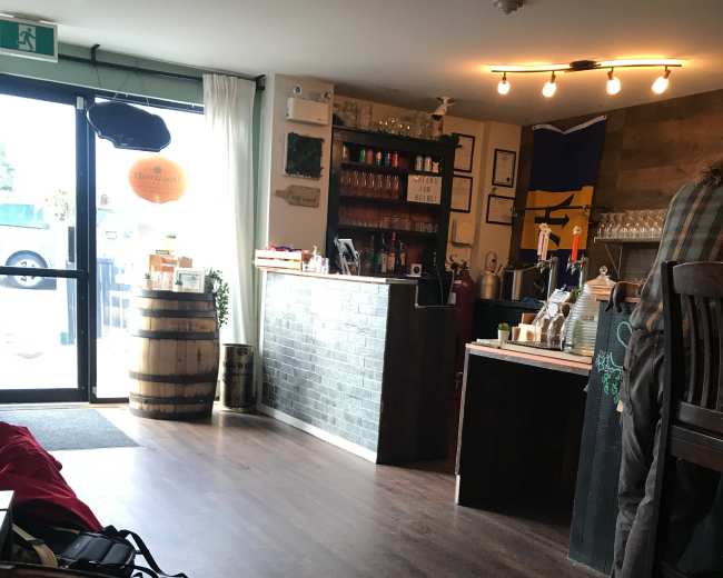 The image shows the interior of a bar or café with wooden flooring, a counter lined with bottles, and large windows letting in natural light.