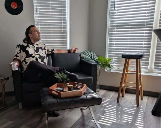 A man sits on a black couch in a well-lit room, holding a mug and looking thoughtfully towards a window with blinds.