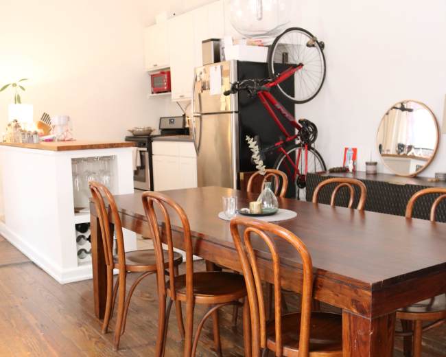 A long wooden dining table with several chairs is positioned in a room featuring a kitchen area with modern appliances and a bicycle hanging on the wall.