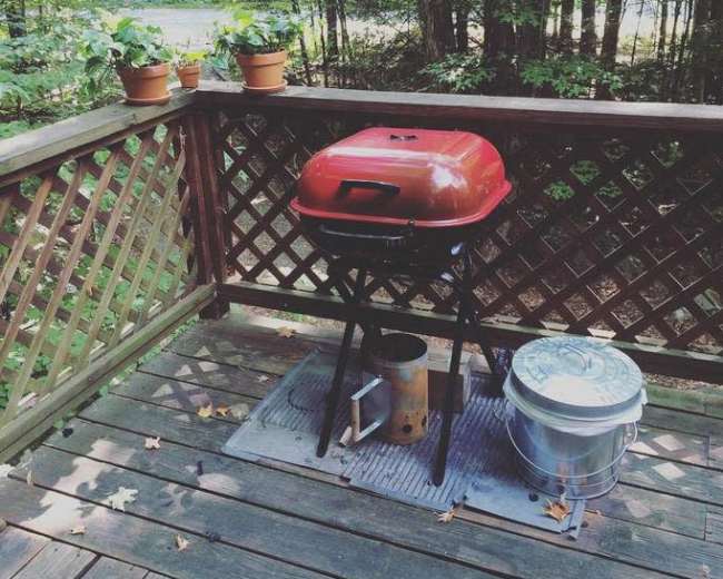 A red grill is positioned on a wooden deck surrounded by green trees, with potted plants nearby.
