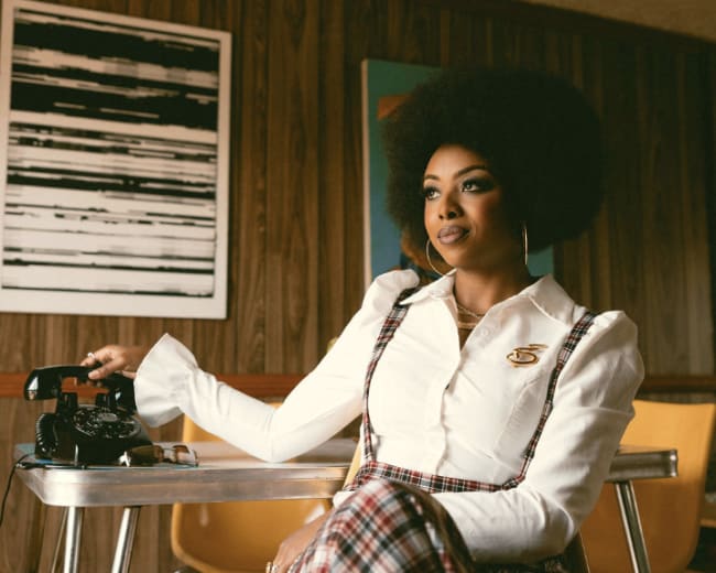 A woman with an afro hairstyle sits at a table in a retro-style room with wooden paneling, featuring a vintage telephone and a chandelier above.