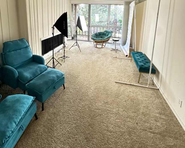 The image shows a spacious room with a light-colored carpet, teal armchairs, a green lounge chair, and several photography lighting equipment set up near large windows.
