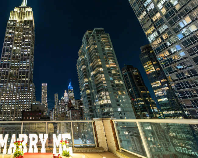 A romantic rooftop proposal scene features large light-up letters spelling "MARRY ME," with the Empire State Building illuminated in the background against a night skyline.