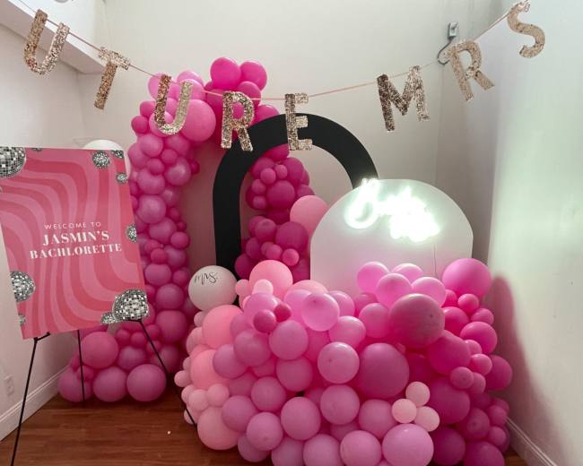 The image shows a festive decoration featuring pink balloons arranged in clusters alongside a sign that reads "Welcome to Jasmin's Bachelorette" and another illuminated sign that says "Mrs."