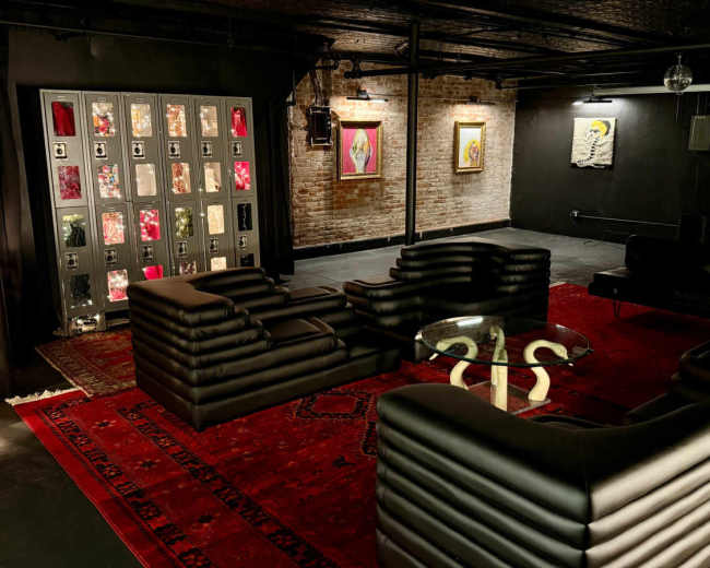 The scene shows a modern, minimalist lounge area featuring black leather seating arranged around a glass coffee table, with art pieces displayed on the walls and a patterned rug on the floor.