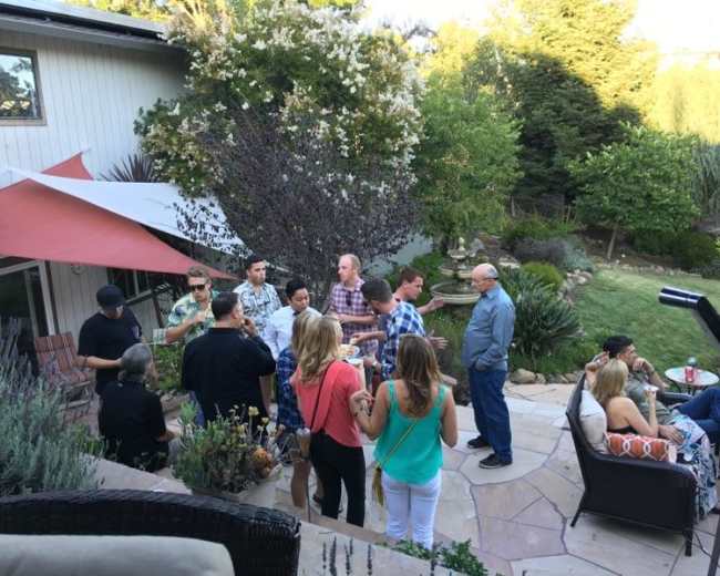 A group of people socializes outdoors in a backyard setting, surrounded by greenery and landscaping.