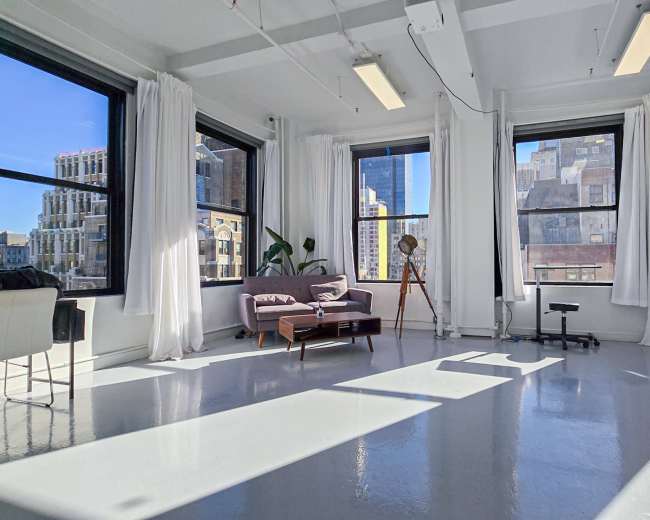 The image shows a bright, spacious room with large windows, featuring a purple sofa, a wooden tripod, and white curtains, set against an urban skyline.