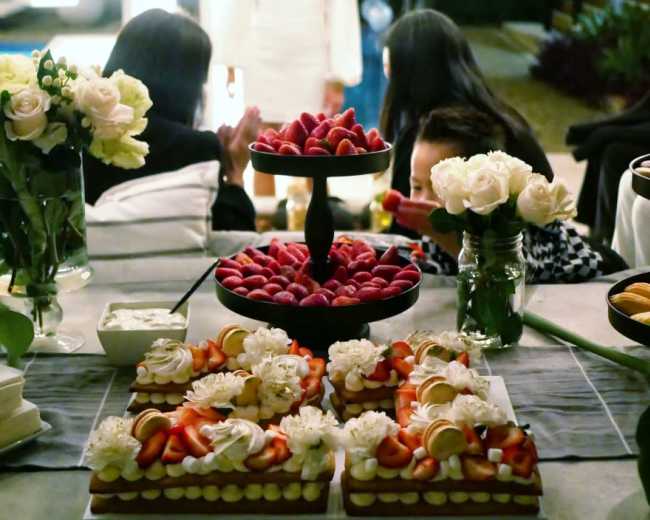 A table displays an assortment of cakes and fruit in the foreground, while a woman in a white dress stands in the background against a backdrop of festive lights and decorations.
