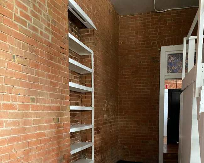 The image shows a room with exposed brick walls, a gray floor, and white shelves mounted on the left wall.