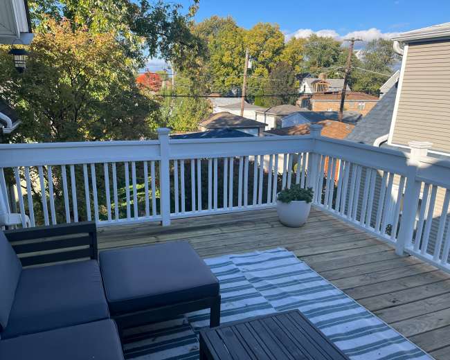 A wooden deck with seating overlooks a neighborhood filled with trees and rooftops under a clear blue sky.