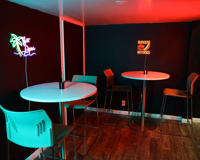 The image shows a small indoor space with two high tables and chairs, illuminated by neon signs that read "Your Space" and "BURGERS HOTDOGS."