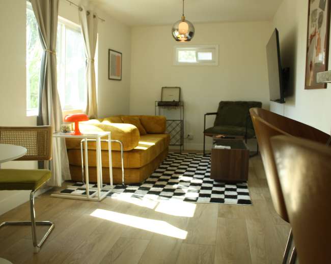 The image shows a modern living room with a yellow sofa, a green chair, a round table, and large windows allowing natural light to enter.