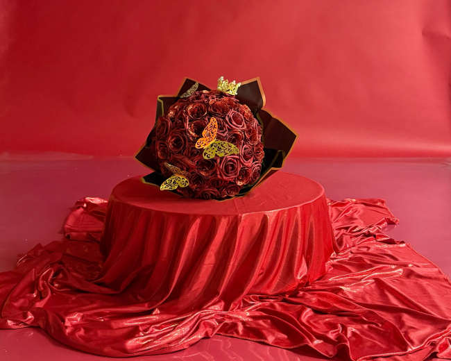 A bouquet of red roses arranged in a circular shape sits on a round table covered with red fabric, all against a red backdrop.