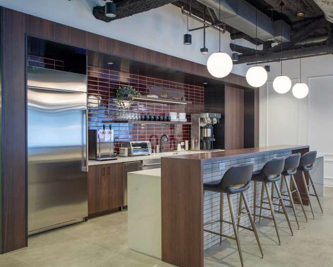 A modern office kitchen area featuring a stainless steel refrigerator, a coffee machine, and a bar with four high stools under pendant lighting.