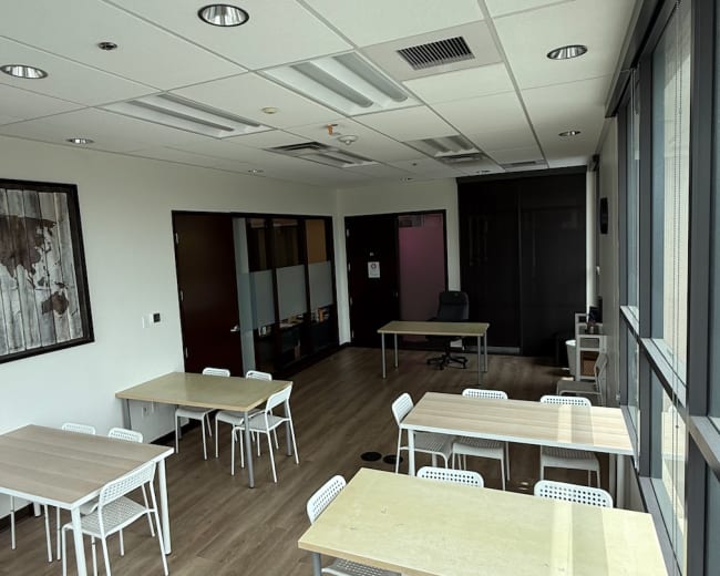 The image shows a modern meeting room with several tables and chairs arranged, large windows allowing natural light, and a dark partitioned area in the background.