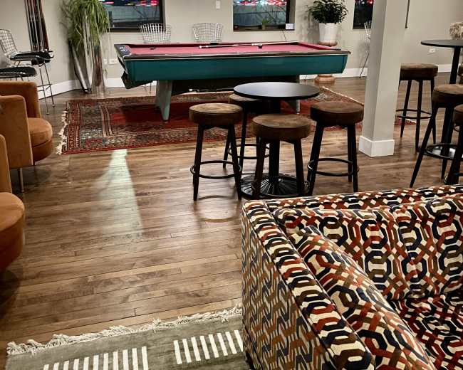 The image shows a modern lounge area with a pool table in the center, surrounded by various seating options and decorative plants.