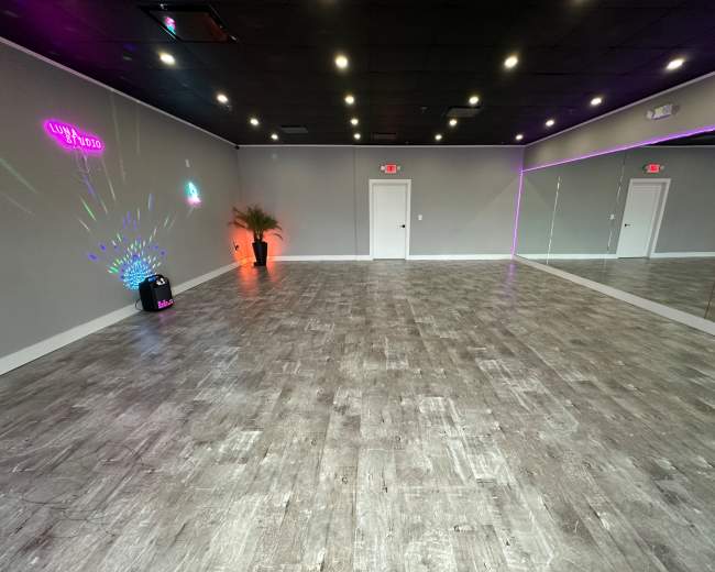 The image shows a spacious, empty room with a wooden floor, gray walls, and mirrored panels, illuminated by various lights.