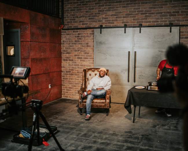 A person sits in an armchair in front of a large sliding door, while a figure in the background prepares food on a table.