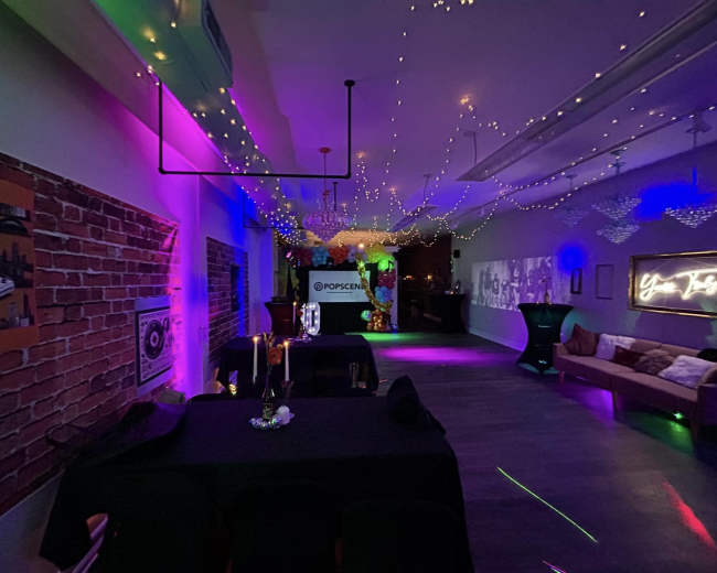 The image shows a dimly lit event space decorated with string lights and neon colors, featuring tables and a large projection screen.