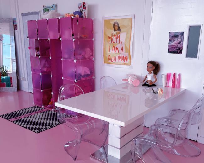 A bright pink room features a white table surrounded by transparent chairs, with a pink storage unit and a poster on the wall.