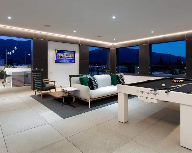 A modern lounge area featuring a billiard table, a sectional sofa, and large windows with a view of mountains at dusk.