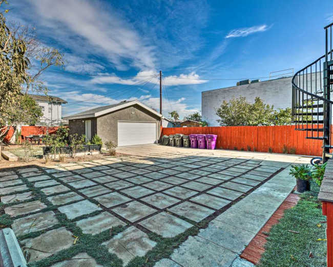 The image shows a spacious backyard with a stone patio, a garage with a white door, and colorful trash bins behind a wooden fence.