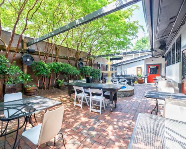 The image shows an outdoor patio area featuring brick flooring, tables, and greenery, with a fountain in the center.
