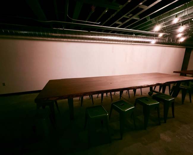 A long wooden table with green stools is set up in a dimly lit room with exposed ductwork and a blank wall.