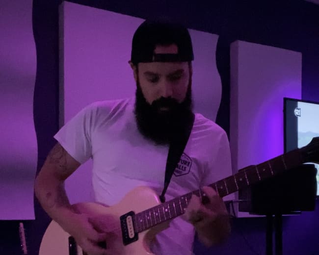 A man with a beard plays an electric guitar in a dimly lit room with purple lighting.