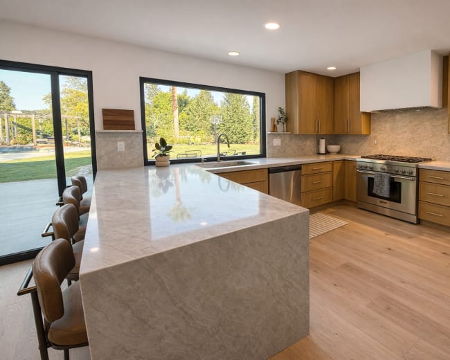 The image shows a modern kitchen with wooden cabinetry, a large island with a marble countertop, stainless steel appliances, and large windows overlooking an outdoor space.