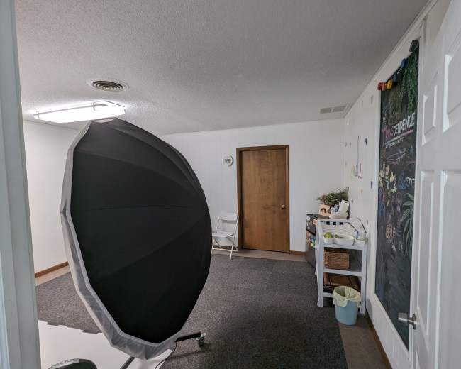 The image shows a well-lit room featuring a large black umbrella-style light softbox, a closed wooden door, and a decorated wall with colorful letters.